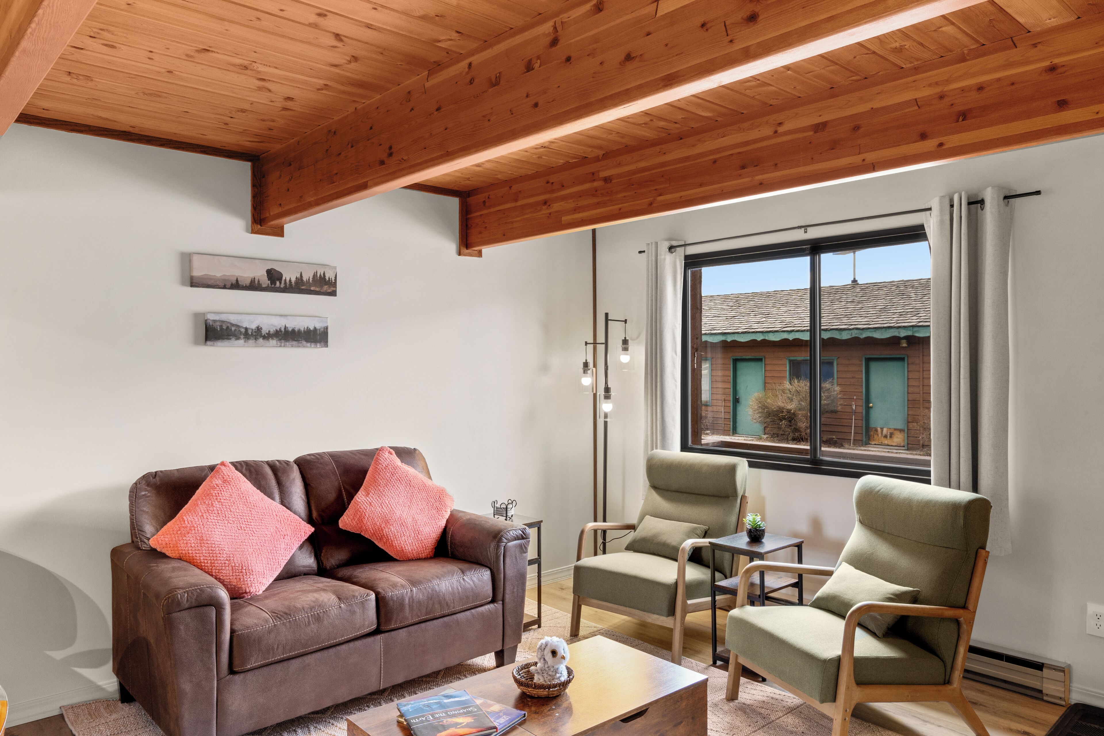 A living room with a brown leather sofa, two green armchairs, a wooden coffee table, and a large window with a view of a building.
