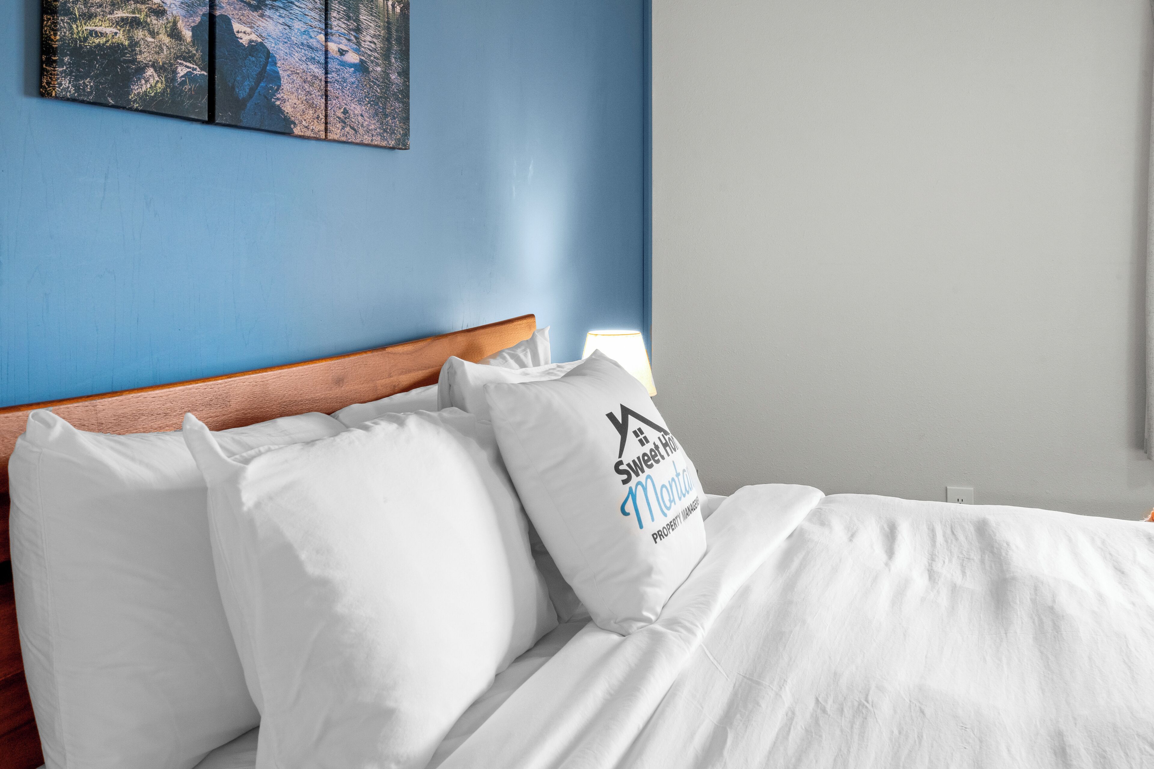 Un lit bien fait, avec une literie et des oreillers blancs, une tête de lit en bois et un mur agrémenté d’une touche de bleu et d’un tableau encadré.