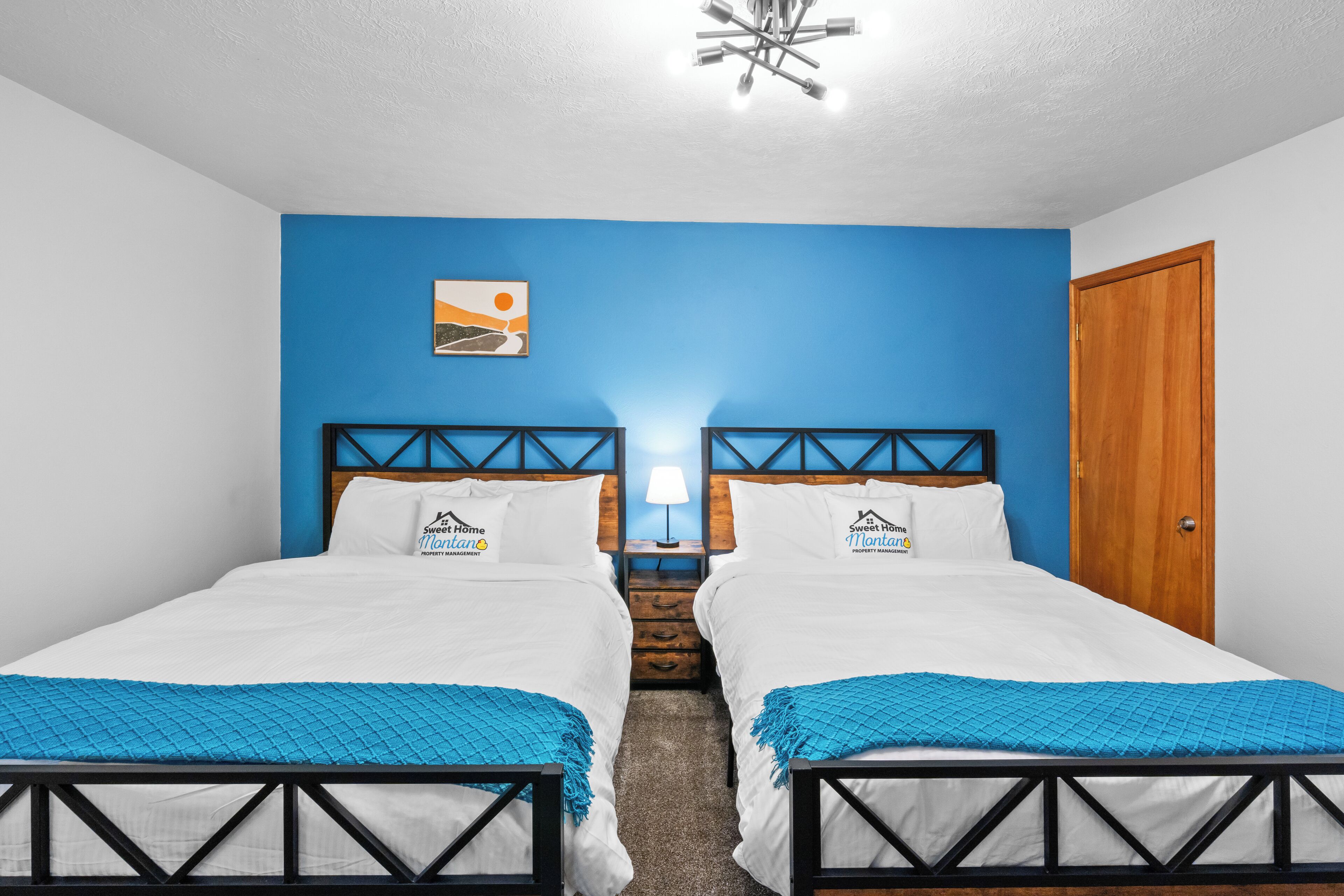 A double room with two beds, a blue accent wall, a wooden door, and a ceiling light fixture.