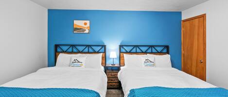 A double room with two beds, a blue accent wall, a wooden door, and a ceiling light fixture.