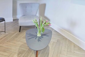 Una habitación moderna con un sillón tapizado de color gris, una mesita redonda con un florero y suelo de madera.