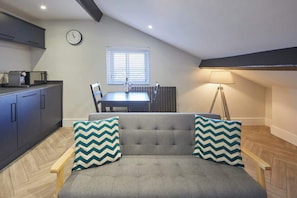 A cozy loft bedroom with a grey sofa, two matching pillows, a dining table, and a clock on the wall.