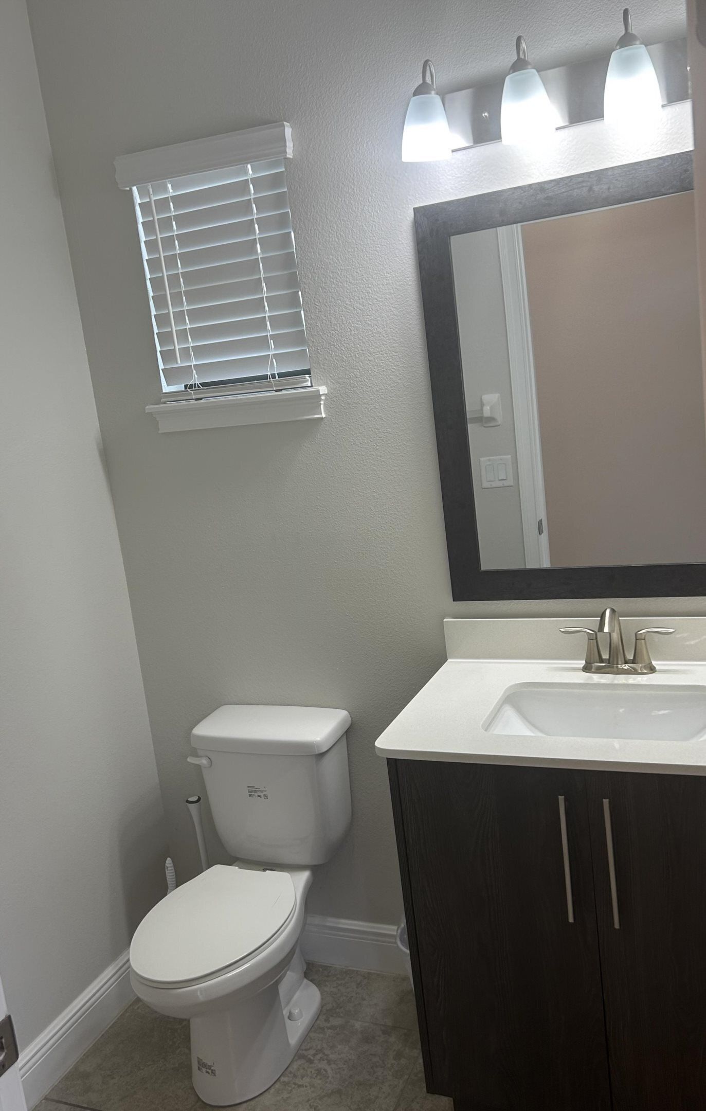 A bathroom with a toilet, a sink, a mirror, and a window with blinds.