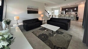 Un salon moderne avec deux canapés noirs, une table basse blanche et un tapis à motifs. Un escalier mène à un espace cuisine doté de meubles sombres et d’un plan de travail blanc.