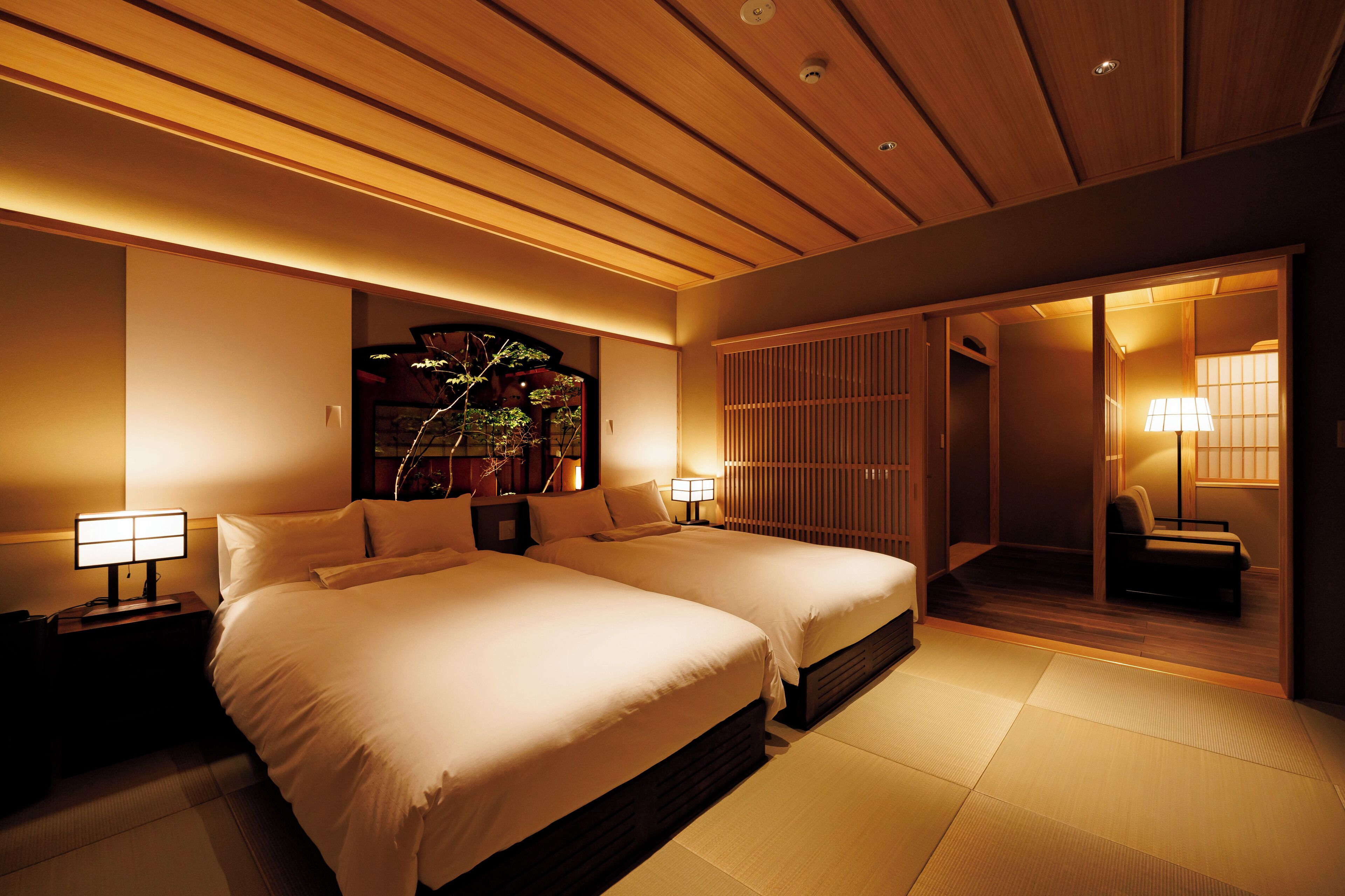 A hotel room with two beds, a nightstand with a lamp, a wall-mounted shelf with decorative items, and a sliding door leading to a sitting area.