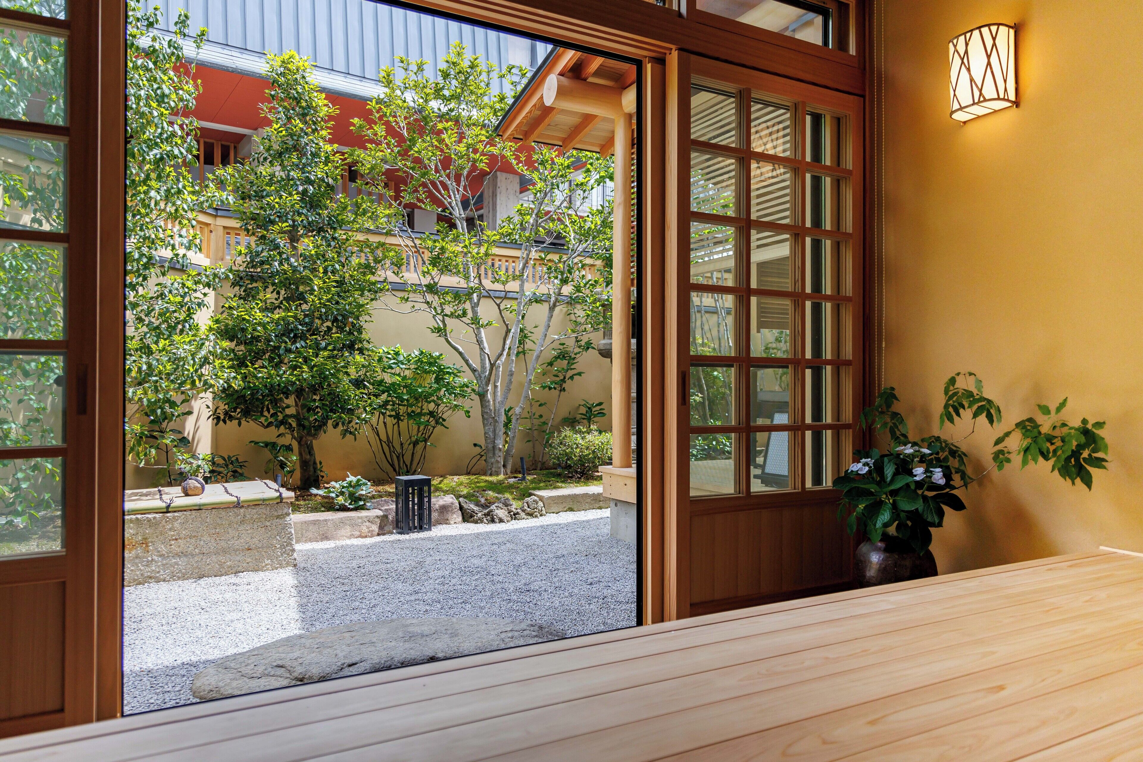 A traditional Japanese-style room with tatami flooring, a wooden sliding door, and a view of a garden with a tree and a building in the background.