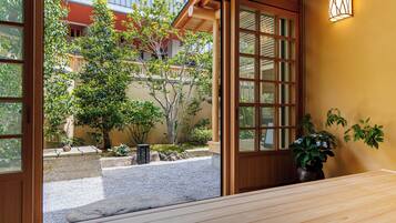 A traditional Japanese-style room with tatami flooring, a wooden sliding door, and a view of a garden with a tree and a building in the background.
