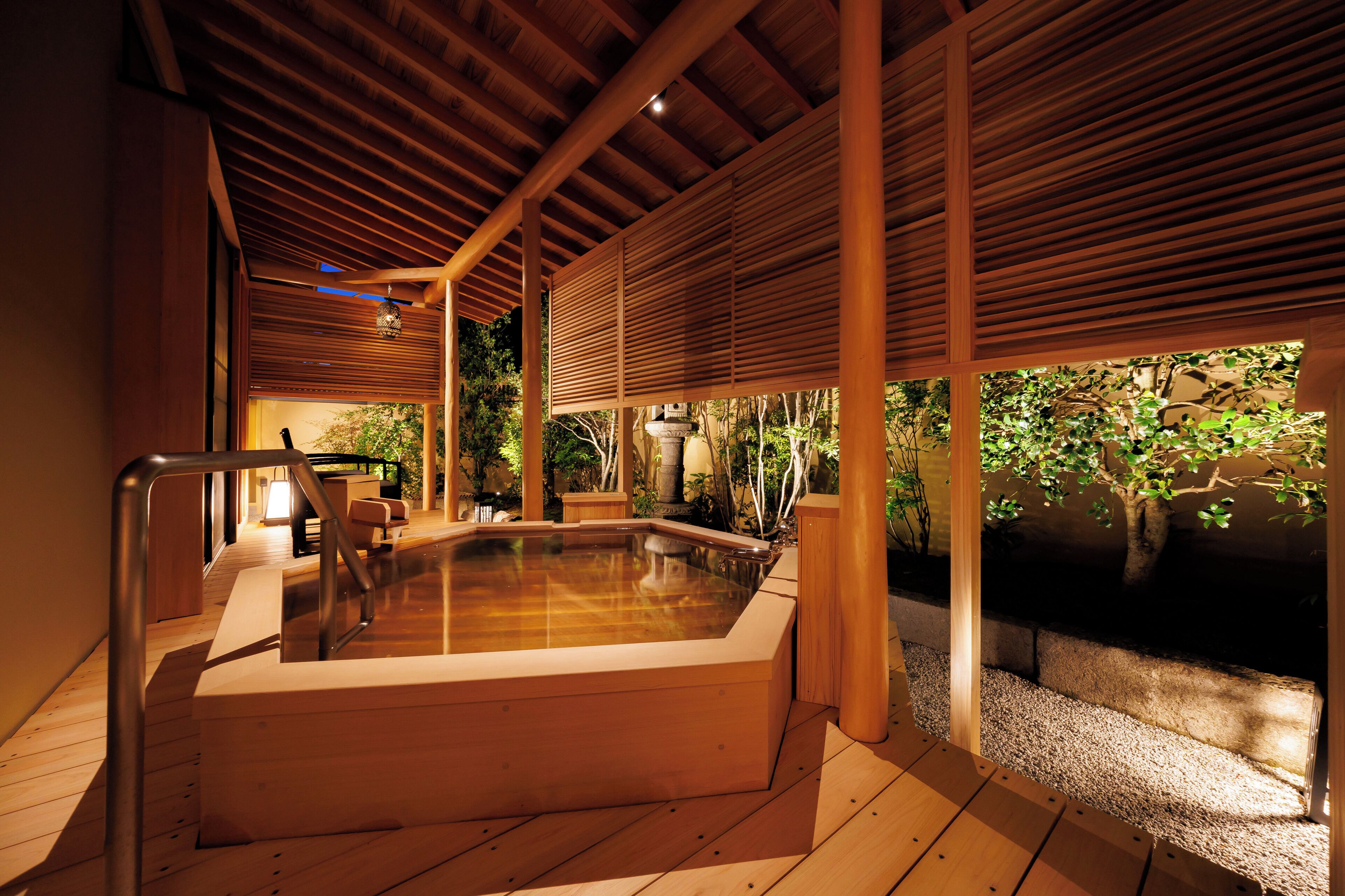 Banheira de hidromassagem sob uma pérgola de madeira, com vista para árvores e uma construção de pedra.