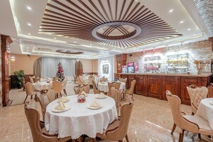 Uma área de refeições de hotel com mesas redondas, cadeiras bege estofadas e um bar central com parede de pedra e objetos decorativos.