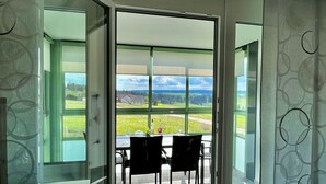 A room with a large glass door leading to an outdoor terrace with a table and chairs, offering a view of a green landscape and distant buildings.
