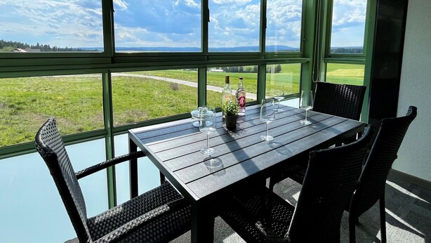 A dining area with a table and chairs, large windows with a view of a green landscape and a road.