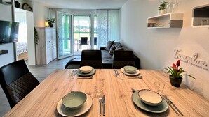 A modern dining area with a wooden table set for two, a sofa in the background, and a view of the outdoors.
