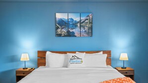 A hotel room with a large bed, two bedside tables with lamps, a wall-mounted painting of a mountain landscape, and a pillow with a logo. - 2BR condo in West Yellowstone, just 1 minute from the park! Owl's Nest (West Yellowstone)