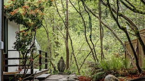 Un sentiero in pietra conduce a un pontile di legno con una statua, circondato da una fitta foresta e una recinzione in legno.