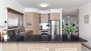 A modern kitchen with a black countertop, wooden cabinets, a stainless steel refrigerator, and a small island with wicker chairs. - `Beach Side` Warrnambool CBD (Warrnambool)
