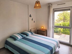 Un dormitorio con cama, mesita de noche, lámpara, ventana con cortinas y vista a zonas verdes.
