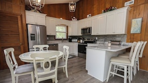 A kitchen with white cabinets, a stainless steel refrigerator, a microwave, and a sink. There is a dining table with chairs and a small island with seating. - Shore 'Nuf - 3 BR Oceanside in Duck (Duck)
