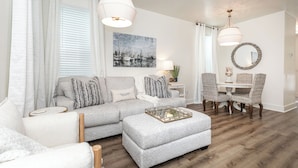 Een woonkamer met een grijze bank, een poef en een eettafel met rieten stoelen. Aan de muur hangt een schilderij en erboven de tafel een ronde spiegel.