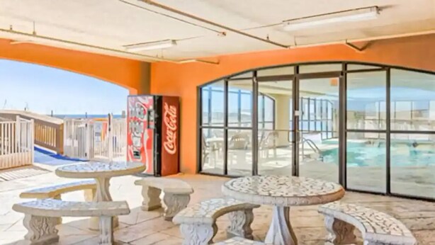 Un espace extérieur aménagé en terrasse, avec des tables et des bancs en pierre, un distributeur automatique de Coca-Cola et une structure vitrée donnant sur une piscine.