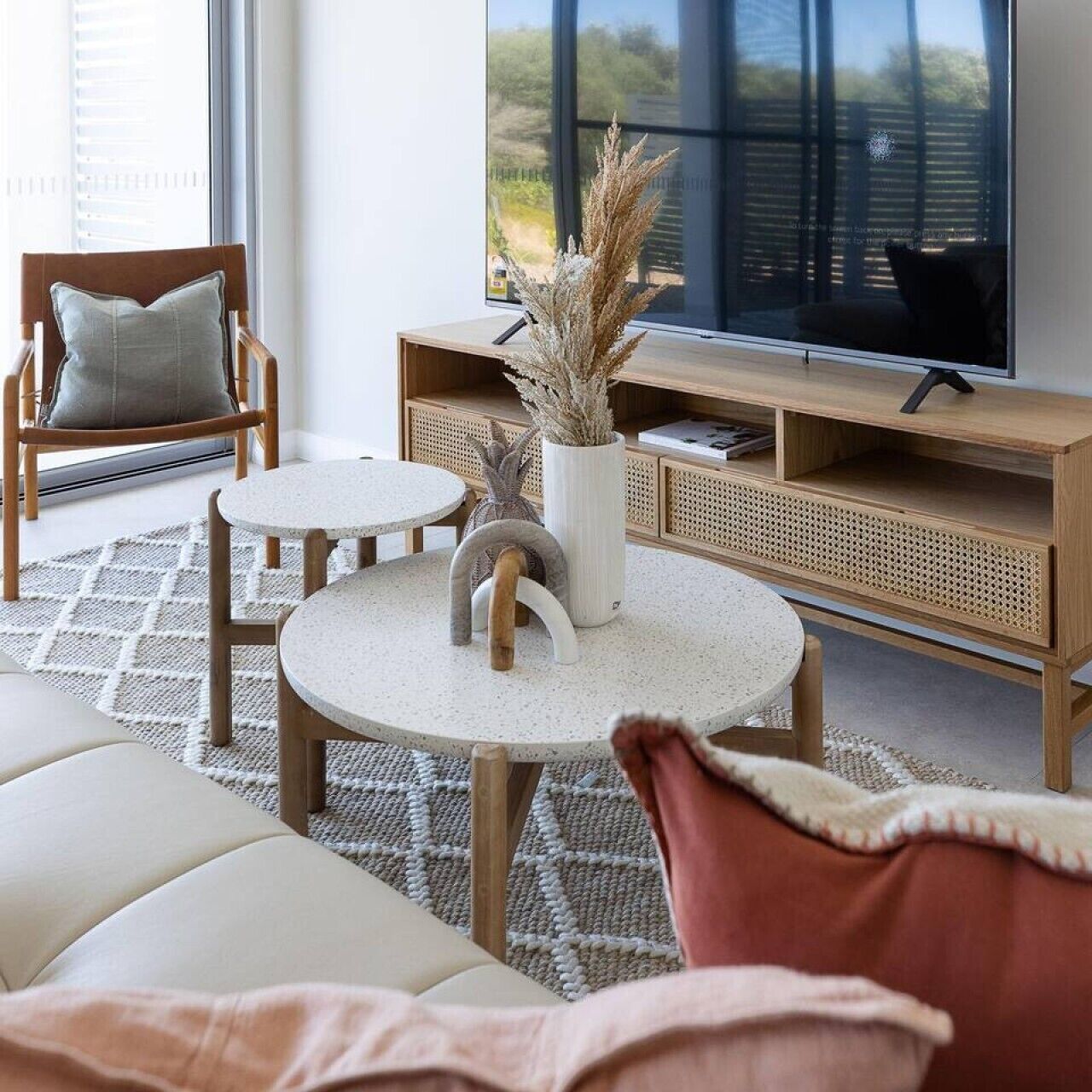 A modern living room with a sofa, two round tables, a TV on a wooden stand, and a large window with a view.