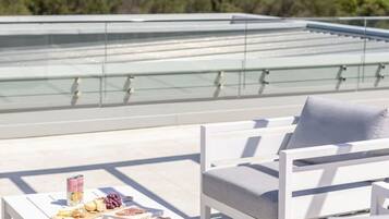 A rooftop terrace with white outdoor furniture, a table with snacks, and a clear view of the ocean.