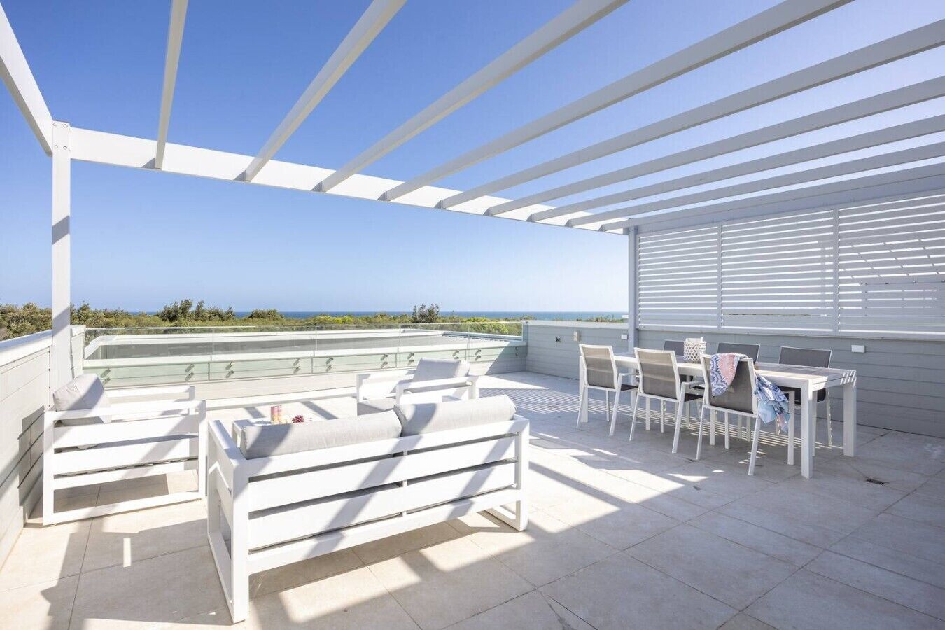 A covered outdoor seating area with white furniture, a table, and a view of the sea.