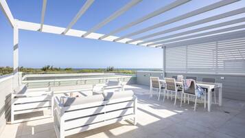 A covered outdoor seating area with white furniture, a table, and a view of the sea.