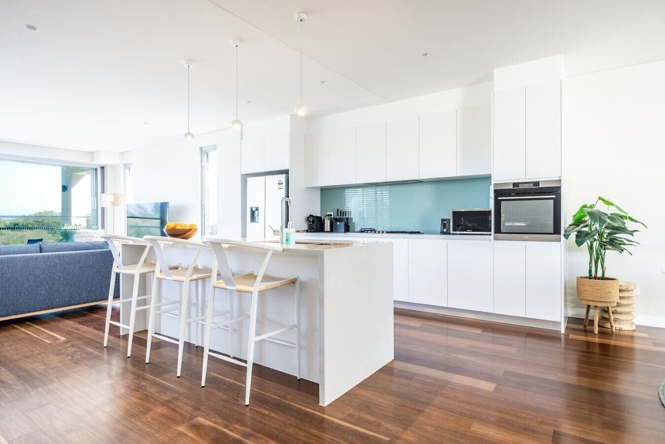 A modern kitchen with white cabinetry, a central island with bar stools, a dining area with a blue sofa, and a large window offering a view.