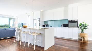 A modern kitchen with white cabinetry, a central island with bar stools, a dining area with a blue sofa, and a large window offering a view.