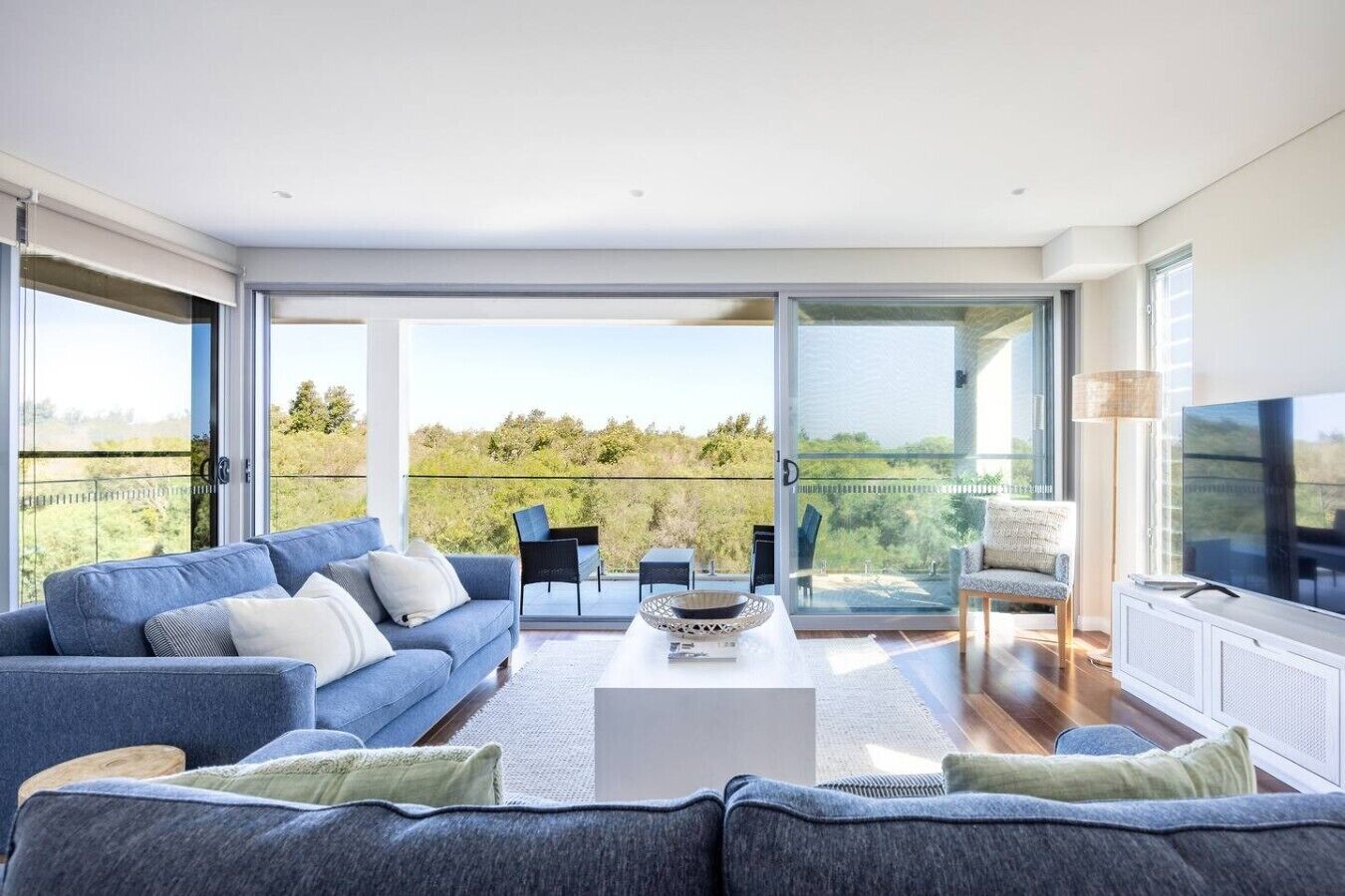 A modern living room with a sofa, coffee table, and chairs, large windows with a view of greenery, and a television.