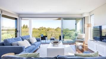 A modern living room with a sofa, coffee table, and chairs, large windows with a view of greenery, and a television.