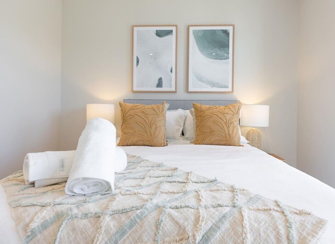 A hotel room with a bed, two framed artworks on the wall, a rolled white towel, and a textured blanket.