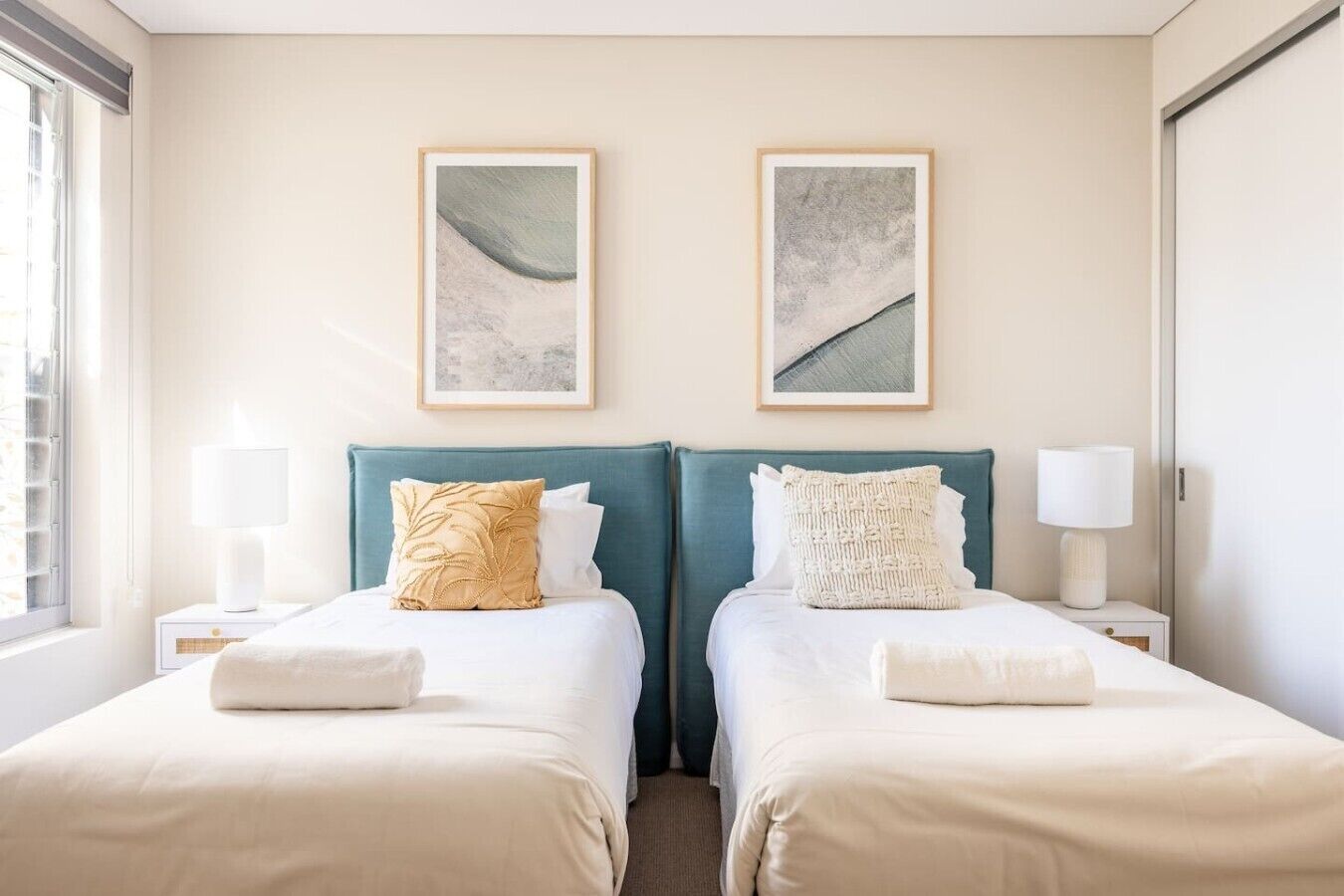 A hotel room with two beds, each with a headboard, a window, and two wall-mounted lamps.