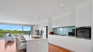 A modern kitchen with white cabinetry, a central island, and a large window overlooking greenery. - Oceanfront Oasis | Stylish 4BR w/ Epic Terrace (Magenta)