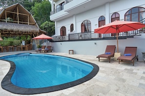 A swimming pool with lounge chairs and umbrellas, a thatched-roof structure, and a white building with arched windows. - Attara Hills Sunset Villa Senggigi (Senggigi)