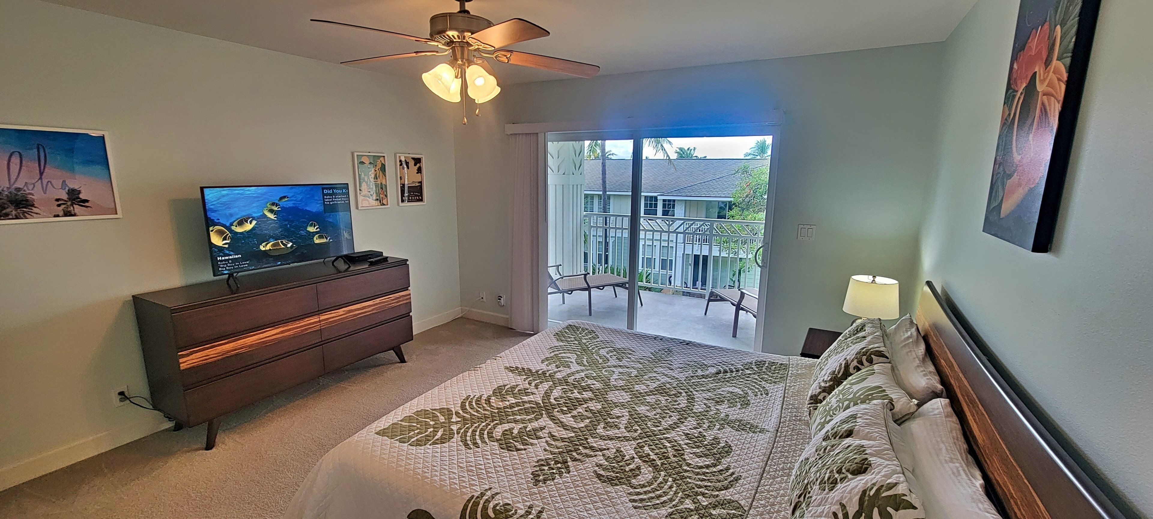 Une chambre à coucher comprenant un lit, une commode, un téléviseur et un balcon avec vue sur une maison et des palmiers.