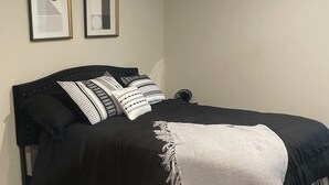 A bedroom with a black bed, white and patterned pillows, a white throw blanket, and two framed abstract artworks on the wall. - Stylish Decor Studio (Tega Cay)
