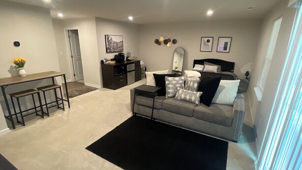 A modern living room with a grey sofa, black and white cushions, a coffee table, a small black table, and a dark-colored rug. There is a dark wooden desk with two stools, a dark-colored cabinet, and a wall-mounted mirror. - Stylish Decor Studio (Tega Cay)