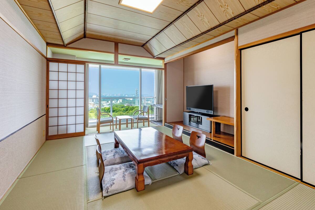 Une pièce de style japonais traditionnel, avec un sol recouvert de tatamis, une table basse avec des coussins, un téléviseur posé sur un meuble en bois et une porte coulissante donnant sur un balcon avec vue sur la ville.