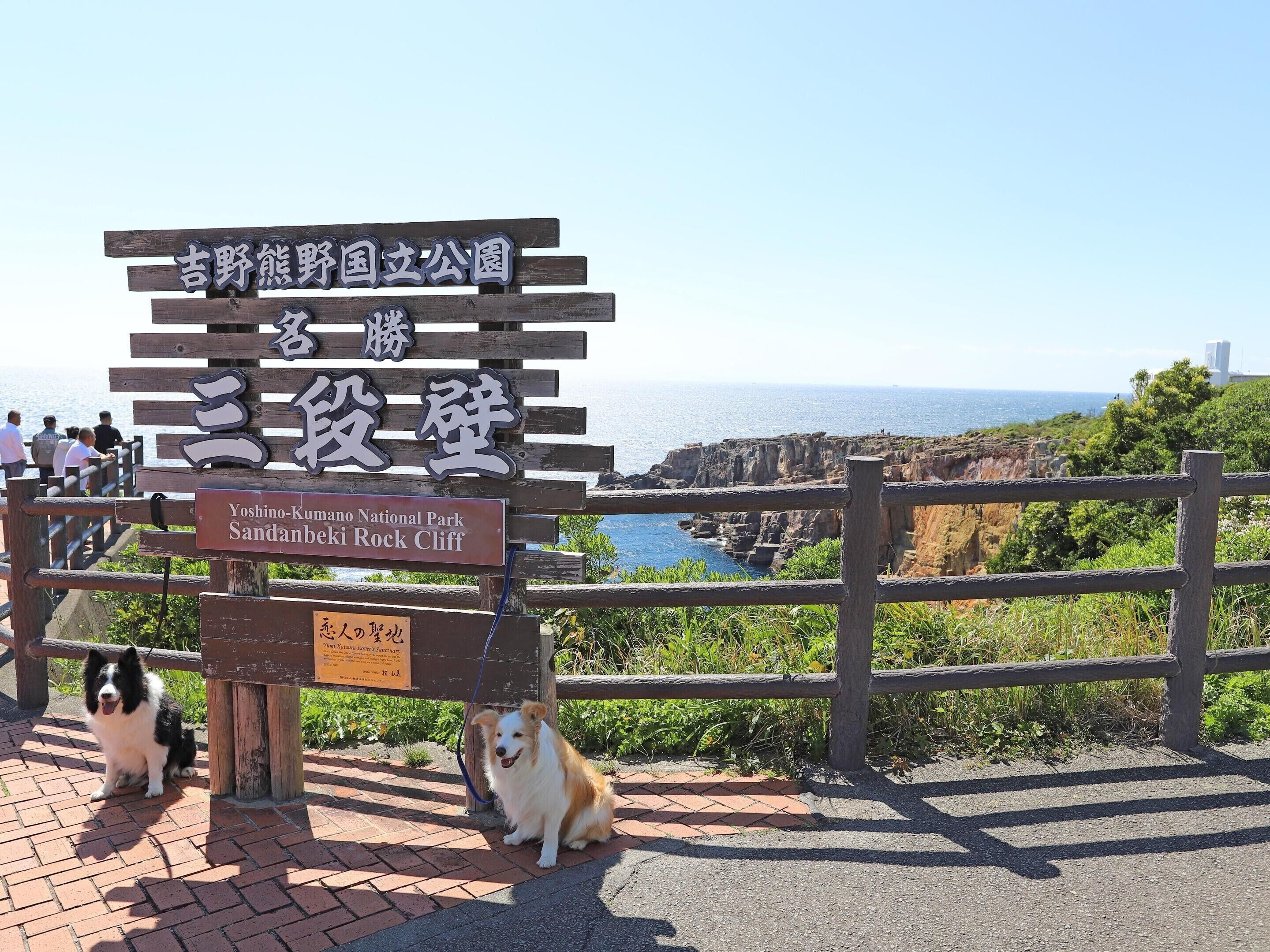 A wooden sign with Japanese text and an English translation for Yoshino-Kumano National Park, indicating a viewpoint of a coastal cliff.