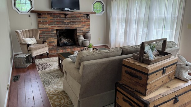 A living room with a brick fireplace, a television mounted above it, a sofa, a chair, and a decorative wooden chest. - Welborn House - Creekside  (Mountain City)