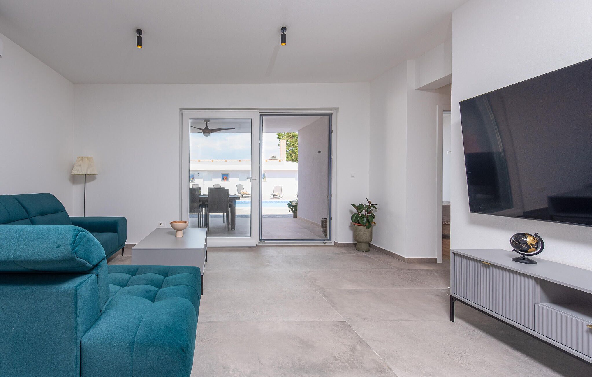A modern living room with teal sofas, a flat-screen TV, and a sliding glass door leading to an outdoor dining area.