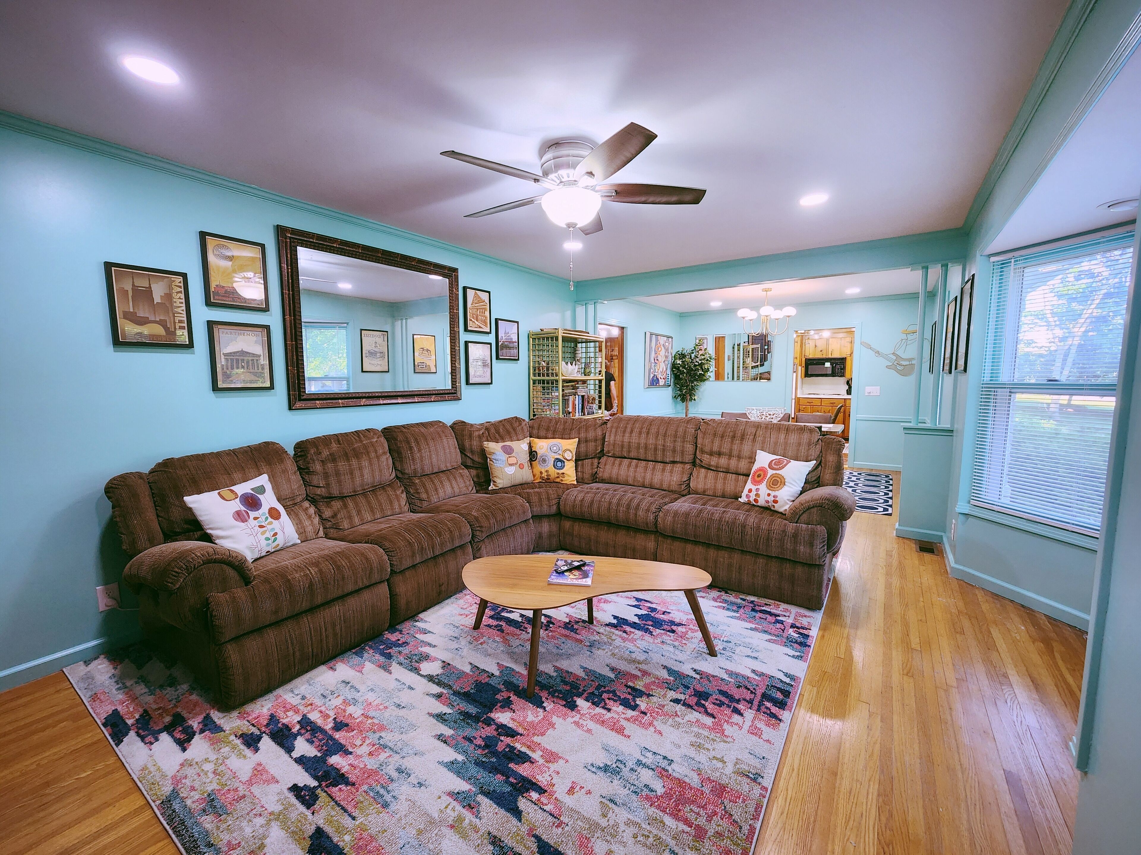 A living room with a brown sectional sofa, a wooden coffee table, and a colorful rug. There is a ceiling fan, a large mirror, and framed pictures on the walls.