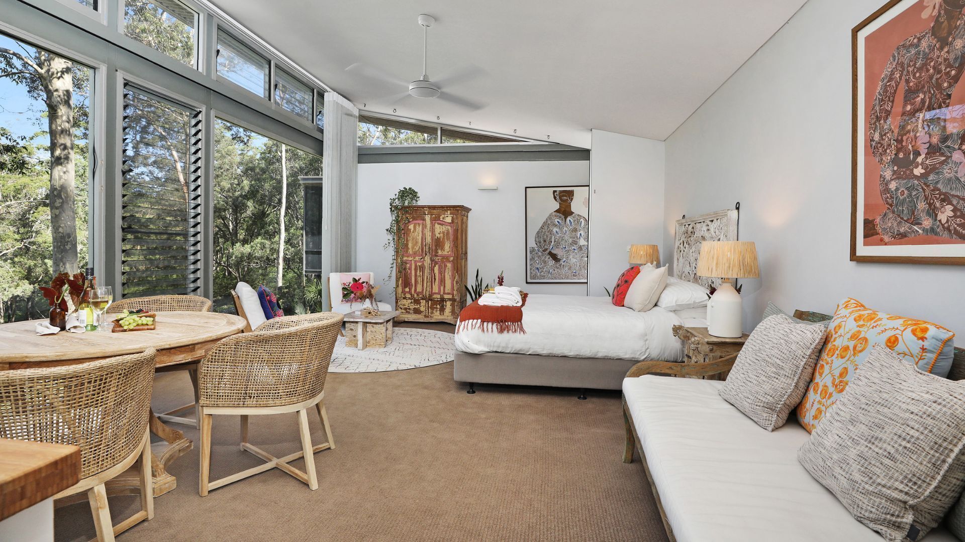 A modern bedroom with a large window, a bed, a sofa, a dining area with wicker furniture, and a vintage cabinet.