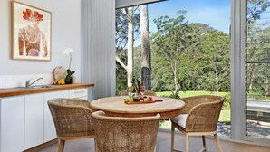 A dining area with a round table, wicker chairs, and a view of a forest through large windows. - Saltwater Creek - immerse yourself in nature (Barringella)