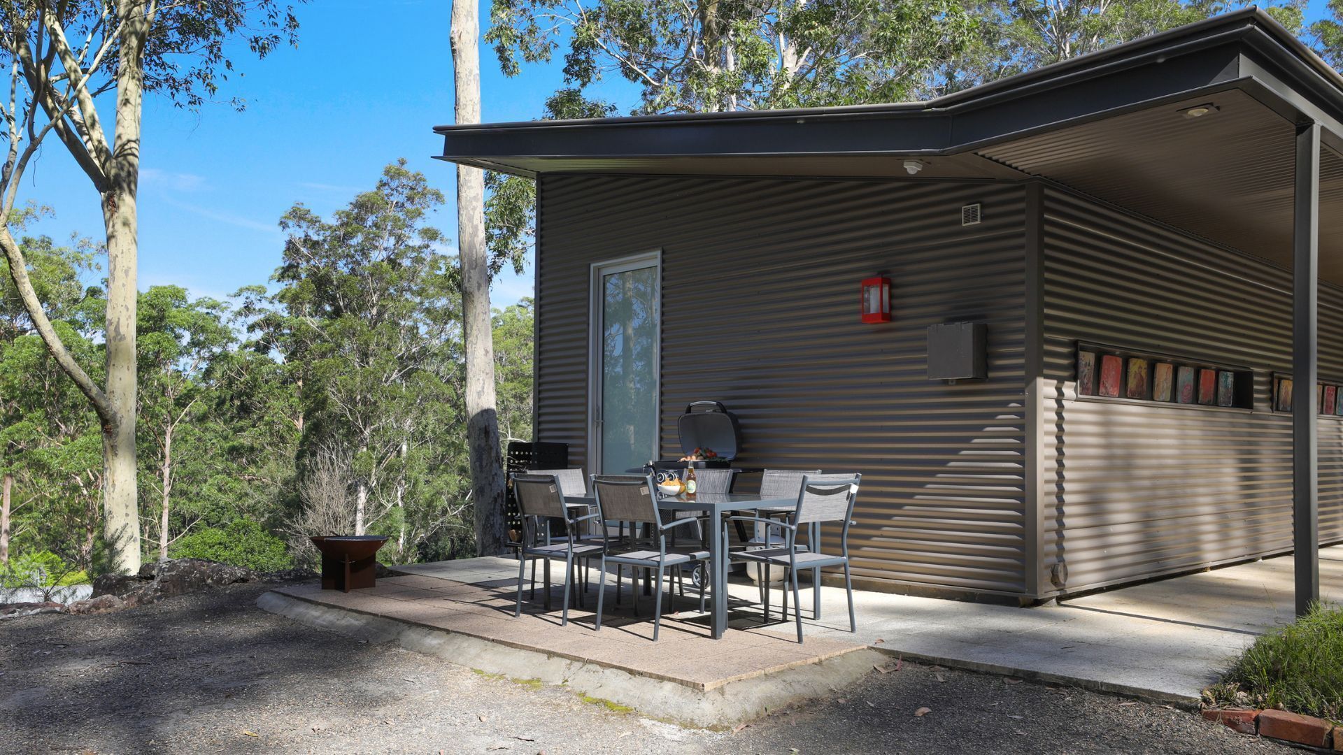 A small, single-story building with a flat roof, a sliding door, and a patio area with a table and chairs.