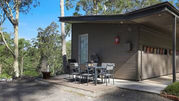 A small, single-story building with a flat roof, a sliding door, and a patio area with a table and chairs.