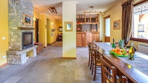 A spacious dining area with a stone fireplace, wooden dining table, and chairs. A kitchen area is visible in the background with wooden cabinets and a hanging light fixture. - Awesome apartment in Pezzoro (Pezzoro)