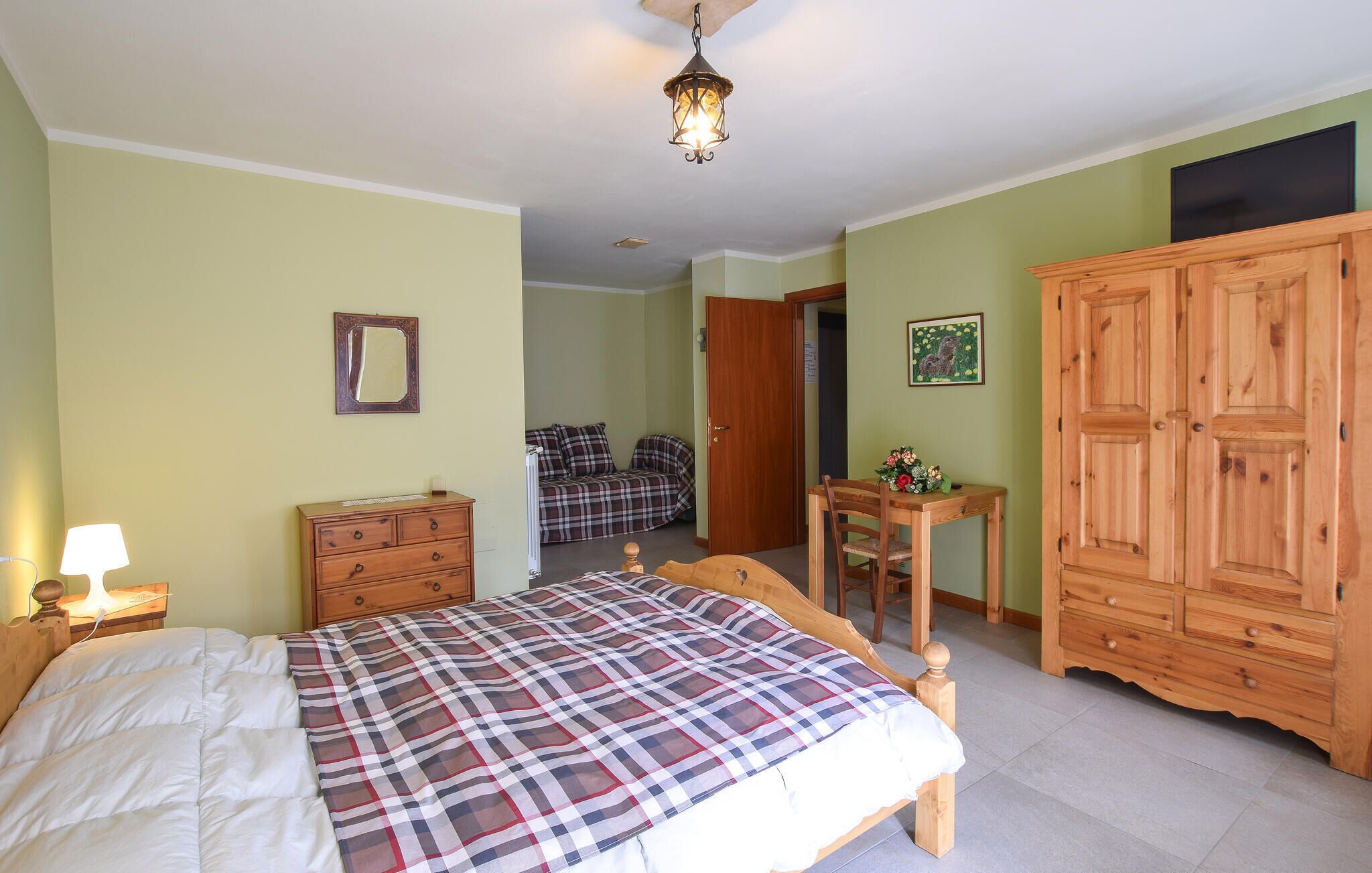 A bedroom with a bed, a wooden dresser, a nightstand with a lamp, a wooden chair, a wooden table with a vase, a door, and a ceiling light fixture.