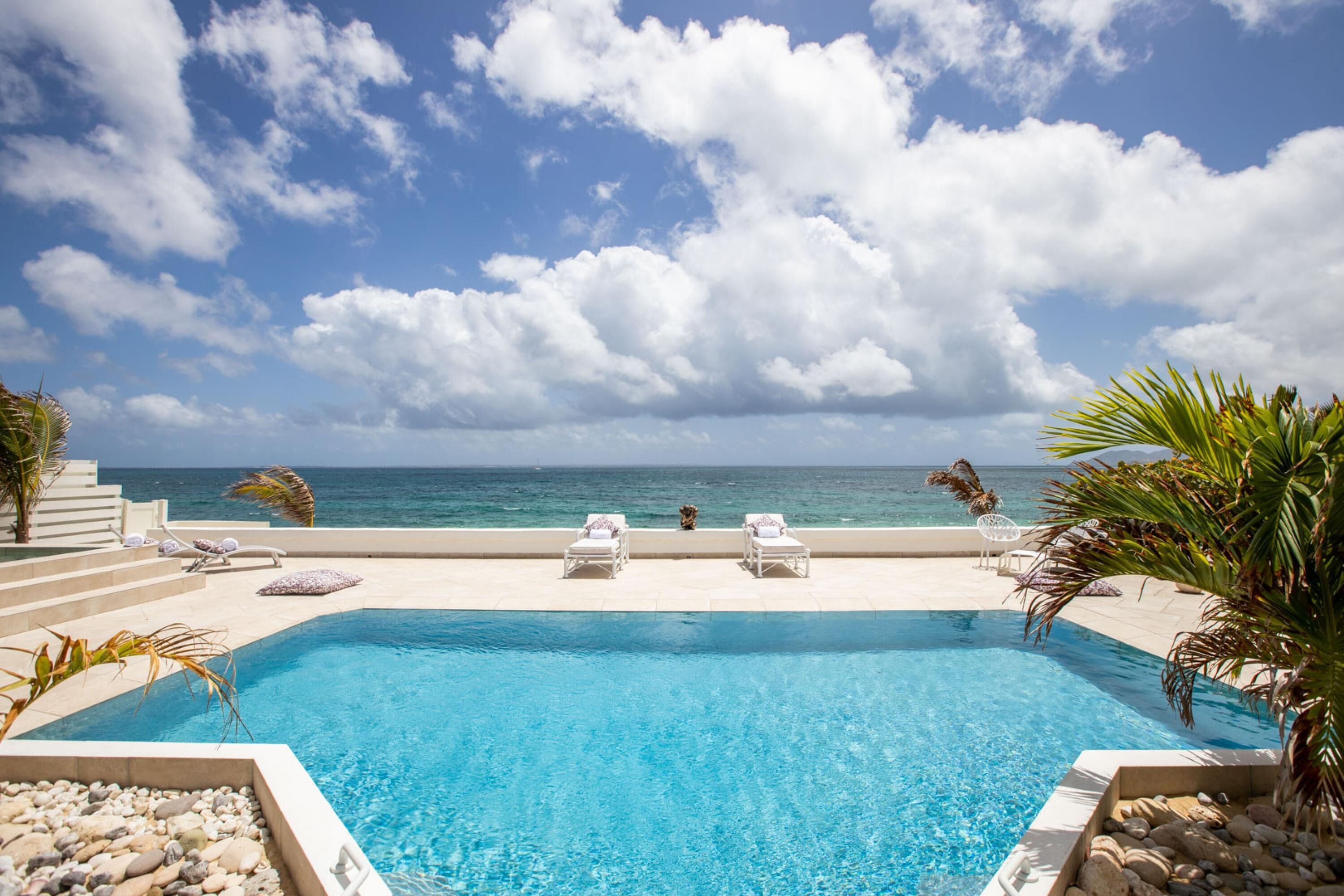 A pool with clear blue water, surrounded by lounge chairs and a palm tree, overlooking the ocean.